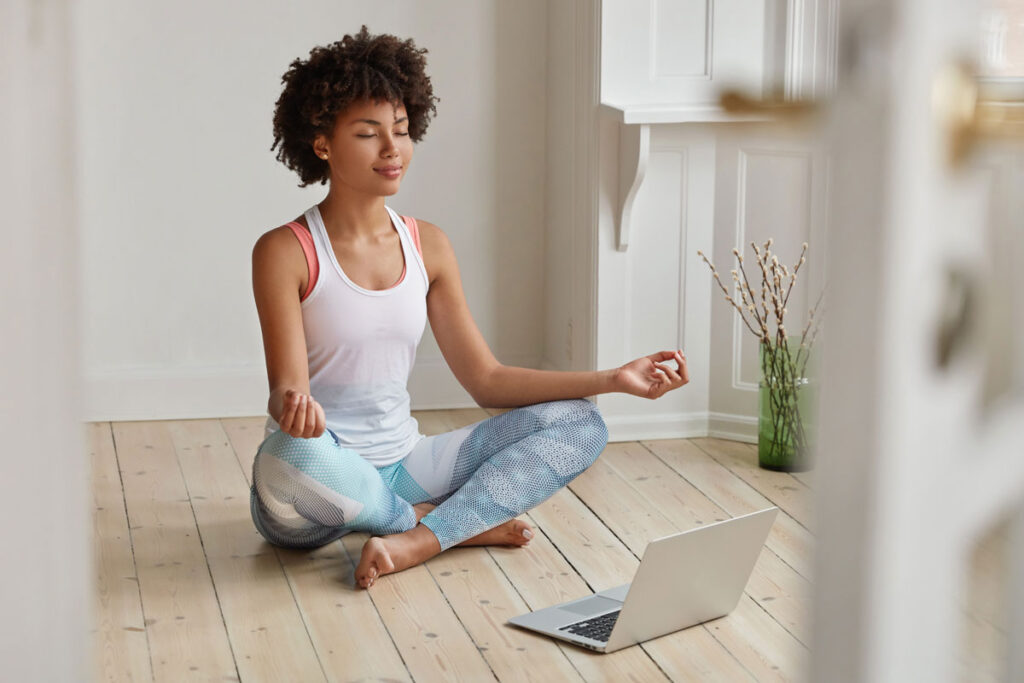 A person sitting cross-legged in a meditation pose on the floor with a laptop open in front of them, participating in a virtual meditation session.